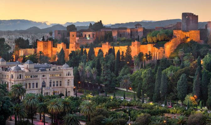 The Alcazaba of Málaga and Gibralfaro Castle illuminated at dusk, overlooking the city, with Málaga City Hall and the park gardens in the foreground.